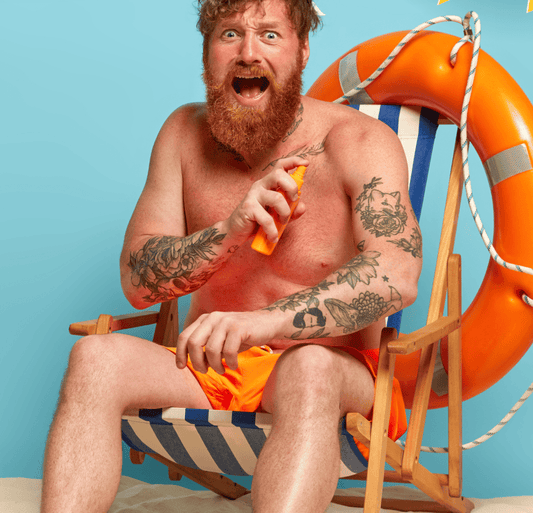 Shirtless man spraying on sunscreen while sitting and making a surprised face.